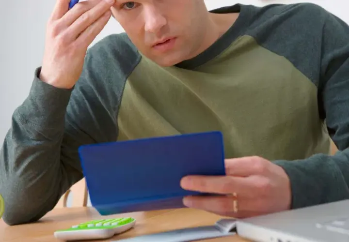 Homme pensif, calculatrice, tasse verte, ordinateur portable. Il se demande combien de temps garder chéquier.