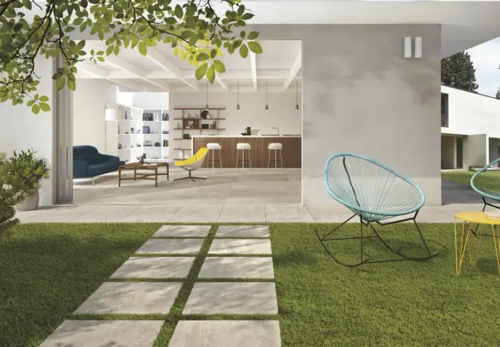 Un sentiero in lastre quadrate su prato verde conduce a un'area living con pavimentazione esterna moderna, che si apre su un interno luminoso con cucina e soggiorno.