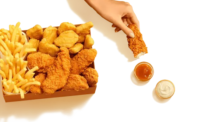 Pudełko McDonald's z kurczakiem i frytkami. Ręka zanurza kawałek kurczaka w sosie. Sprawdź kcal!