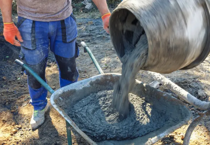 Robotnik w pomarańczowych rękawicach wylewa świeży beton z betoniarki do taczki. Różne rodzaje betonu wymagają różnych metod mieszania i transportu.