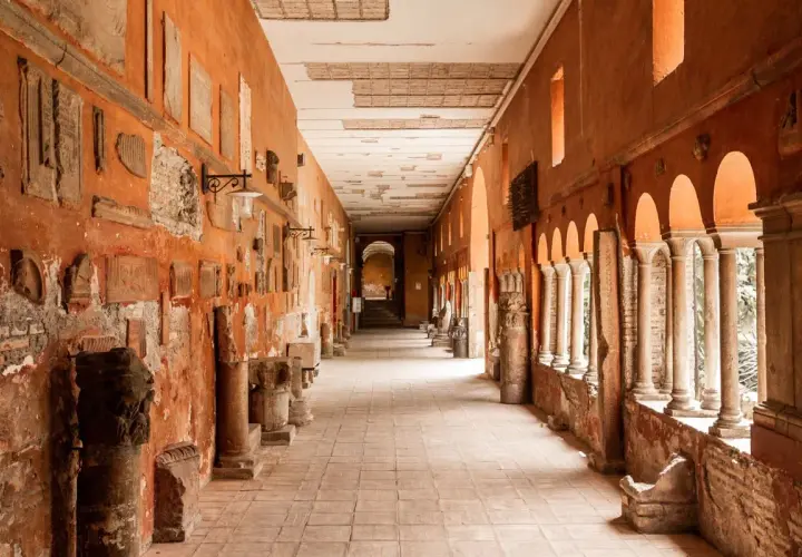 Corridoio con reperti antichi, un'idea di cosa vedere a Trastevere. Colonne, iscrizioni e frammenti decorano le pareti arancioni.