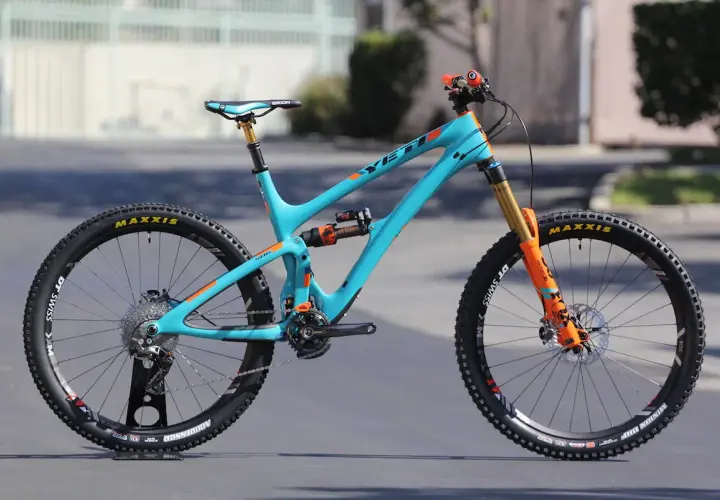 A turquoise Yeti SB6c mountain bike with orange accents, featuring Maxxis tires and Fox suspension, ready for any trail.