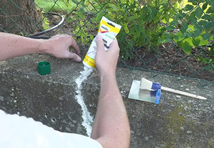 Hände dichten Betonrisse mit einer Tube ab. Ein Spachtel und Pinsel liegen bereit.