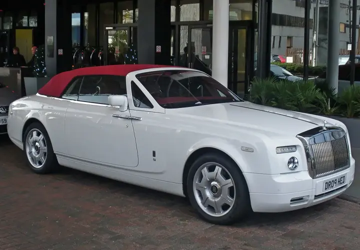 Biały Rolls-Royce Phantom Drophead Coupe z czerwoną, opuszczoną dachem, zaparkowany przed budynkiem.