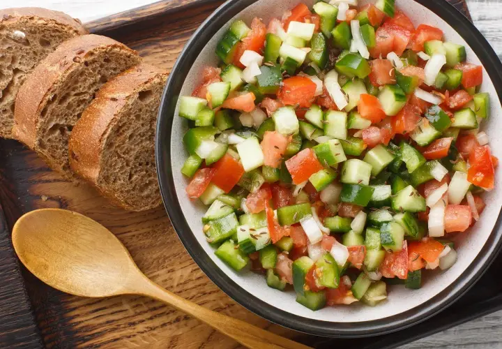 Ensalada pipirrana fresca y colorida, con tomate, pepino, pimiento y cebolla, servida junto a rebanadas de pan integral y una cuchara de madera.