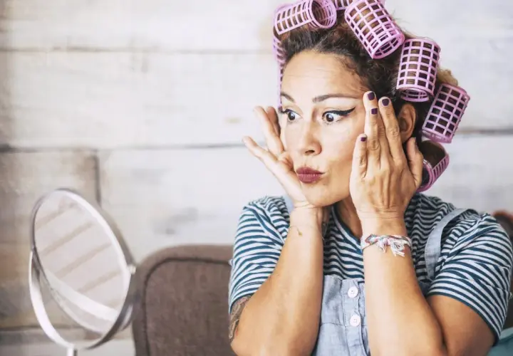Mujer con rulos en el pelo, delineado de ojos y labios pintados, practicando cómo rizar el pelo frente a un espejo.