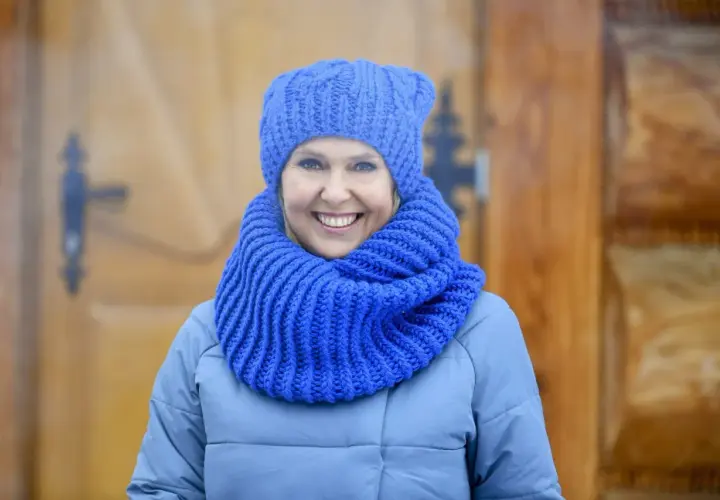 Uśmiechnięta kobieta w niebieskiej czapce i szaliku, idealny przykład, jak dobrać czapkę do płaszcza.