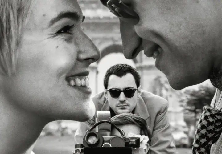 Primo piano di una donna sorridente, con un uomo che la guarda e un altro uomo con occhiali da sole che filma. Un omaggio ai registi importanti.