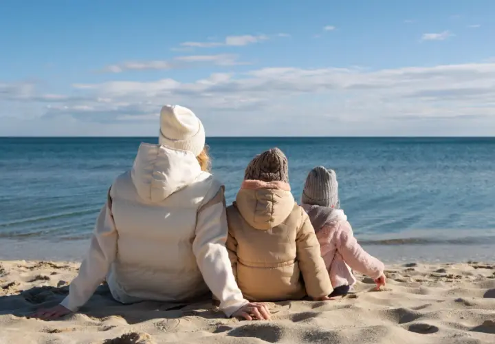 Rodzina w ciepłych ubraniach siedzi na plaży, patrząc na spokojne morze. Pogoda w czerwcu nad morzem bywa kapryśna, ale widok jest piękny.