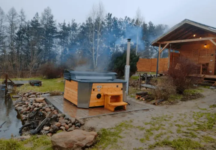 Drewniana balia z parującą wodą, obok której stoi domek w lesie. Idealne miejsce na relaks.