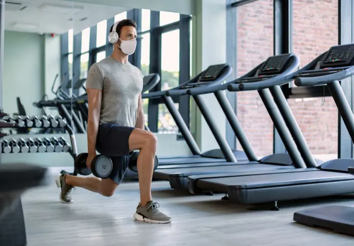 Mężczyzna w masce i słuchawkach wykonuje wykroki z hantlami. To jego trening cardio na siłowni, gdzie dba o formę.