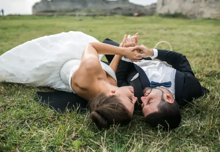 Para młoda leży na trawie podczas sesji plenerowej ślubnej, trzymając się za ręce. W tle ruiny zamku.