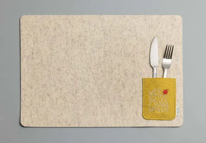 Beiges Platzset aus Filz mit gelber Tasche und der Aufschrift "Wer kleckert muss spülen!". Messer und Gabel stecken darin.