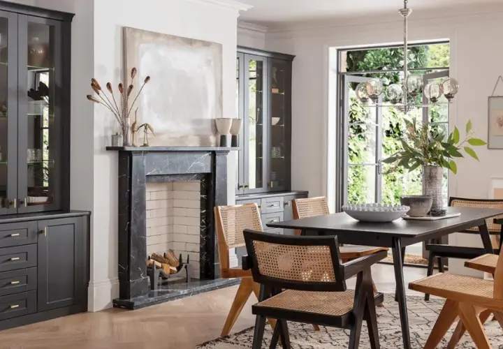 Modern dining room with dark grey cabinets, marble fireplace, and a dark wood table with cane-backed chairs, showcasing current trends in dining room furniture.