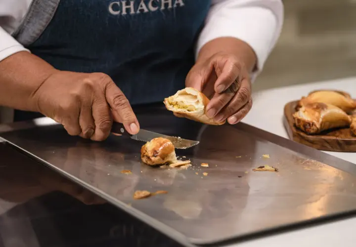 Manos cortan una empanada dorada, revelando su relleno. Otra masa empanada espera en una tabla.