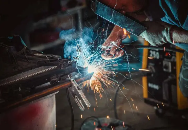 Spawacz w masce tworzy iskrzący łuk, łącząc metalowe elementy. W tle żółta spawarka, a wokół latają iskry.