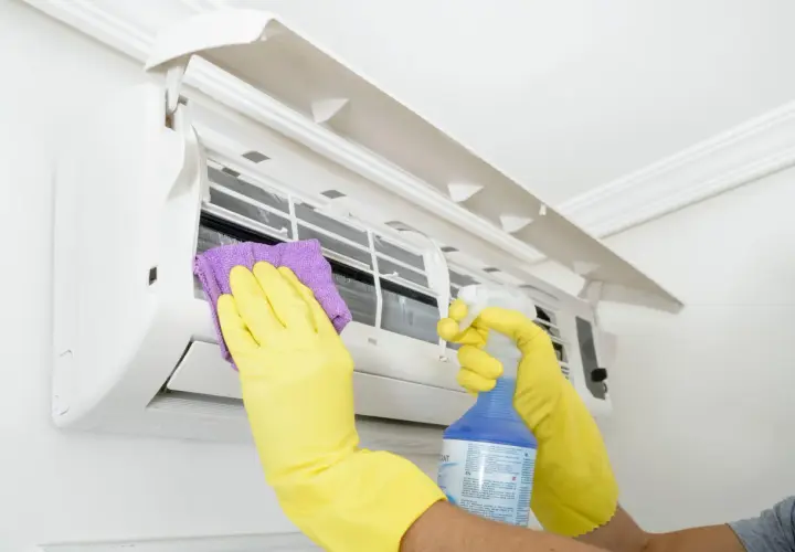 Man con guantes amarillos limpia un aire acondicionado con un paño morado y spray. Aprende como desatascar un aire acondicionado.