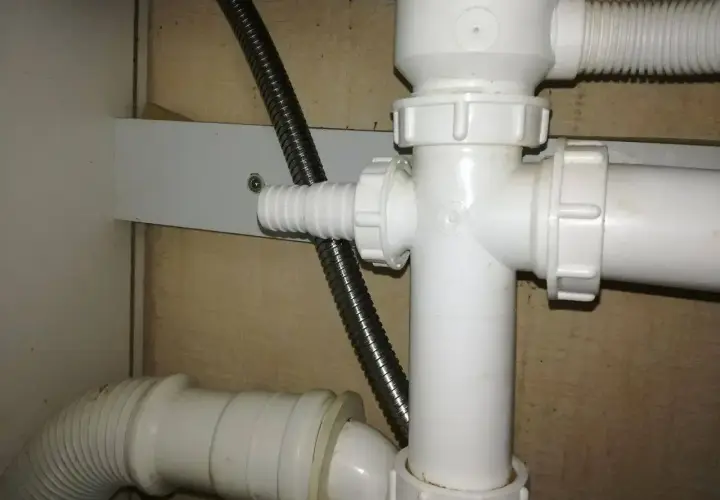 Biały plastikowy trójnik hydrauliczny pod zlewem, do którego podłączona jest elastyczna rura odpływowa i metalowy wąż.