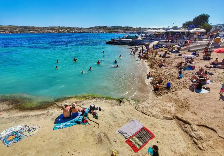 Słoneczna plaża z turkusową wodą, na której kąpią się ludzie, a na piasku odpoczywają plażowicze.