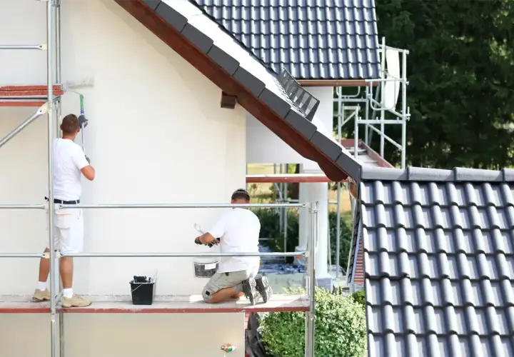Dwóch mężczyzn na rusztowaniu maluje białą elewację. Sprawdzają tynk silikonowy cena za m2 robocizny.