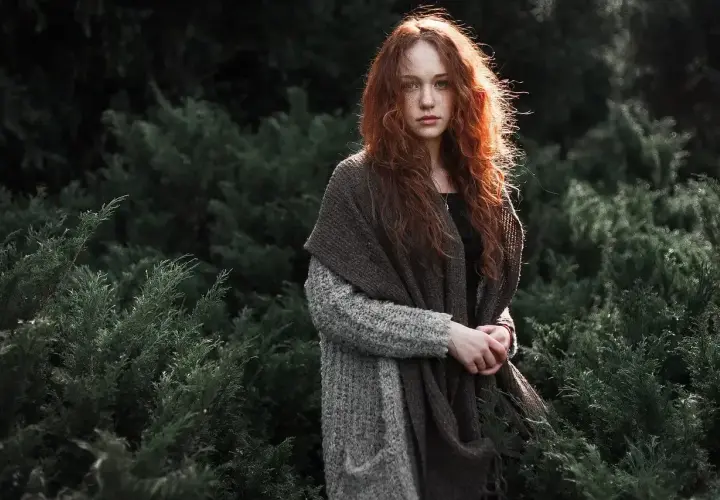 Rudowłosa dziewczyna w swetrze i szalu, otoczona ciemną zielenią. Akt fotografia.