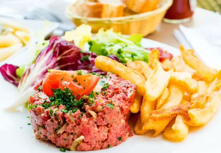 Un délicieux tartare de boeuf, garni d'une tranche de tomate et de persil, servi avec des frites croustillantes et une salade fraîche.