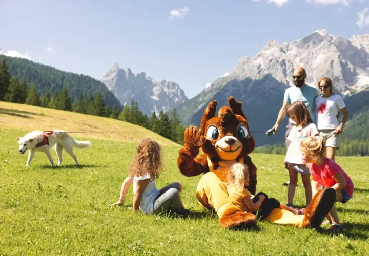 Famiglia al family resort Rainer Sesto, con mascotte e cane su prato verde, montagne sullo sfondo.