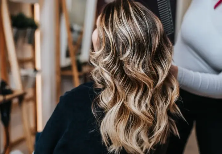 Mujer con cabello castaño y mechas rubias ondulado, visto desde atrás en un salón de belleza.