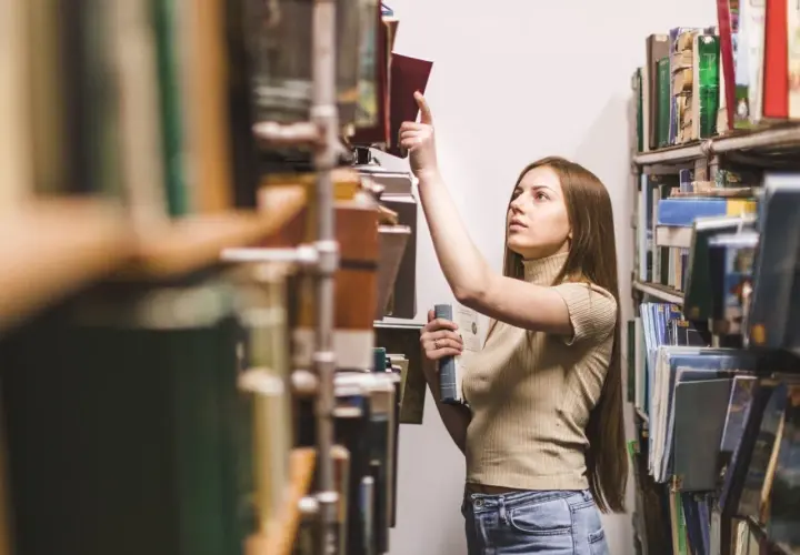 Młoda kobieta w bibliotece szuka inspiracji, zastanawiając się, jakie gatunki literackie są najbardziej poszukiwane przez wydawców w 2026.