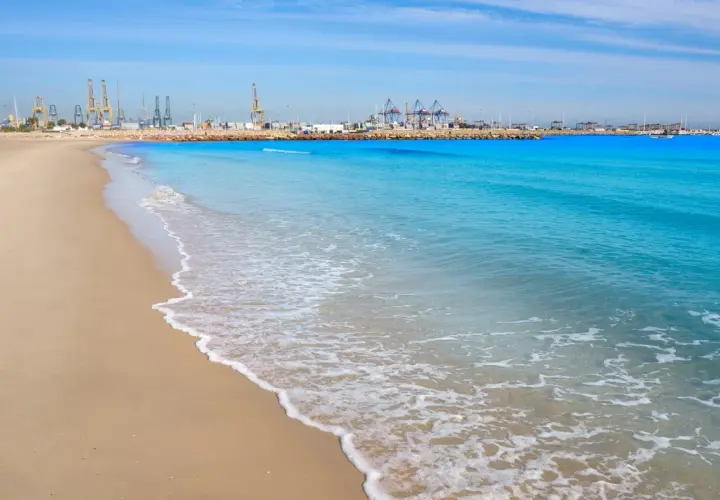 Olas suaves acarician la arena dorada en las **playas de Valencia**, con el puerto industrial de fondo bajo un cielo azul.