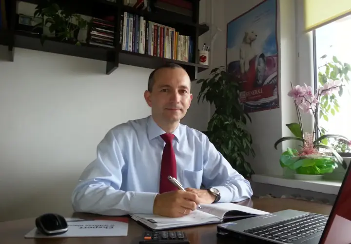 Mężczyzna w jasnej koszuli i czerwonym krawacie siedzi przy biurku, pisząc w notatniku.