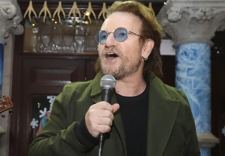 Bono singt ins Mikrofon, trägt eine grüne Jacke und blaue Sonnenbrillen. Der Titel von Bono ist ein bekannter Musiker.
