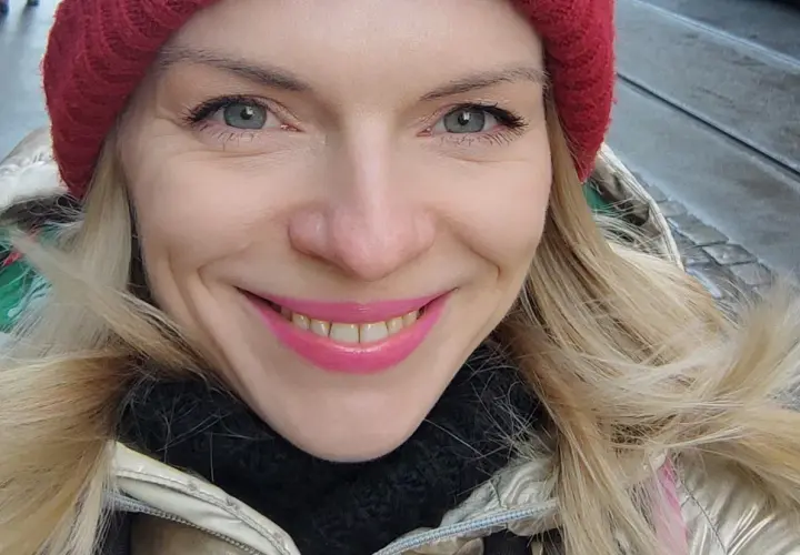 Uśmiechnięta blondynka w czerwonej czapce i złotej kurtce, jakby właśnie spacerowała po ulicach Czech, gdzie ludność cieszy się zimową aurą.