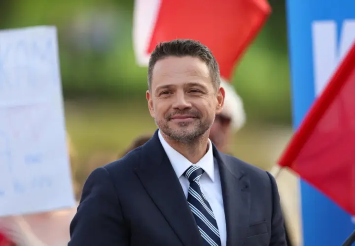 Rafał Trzaskowski w garniturze i krawacie, uśmiecha się na tle flag.