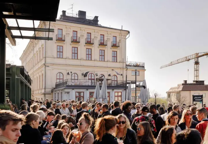 Folk trängs på Mosebacke terassen en solig dag. En stor folkmassa njuter av stämningen framför en gul byggnad.