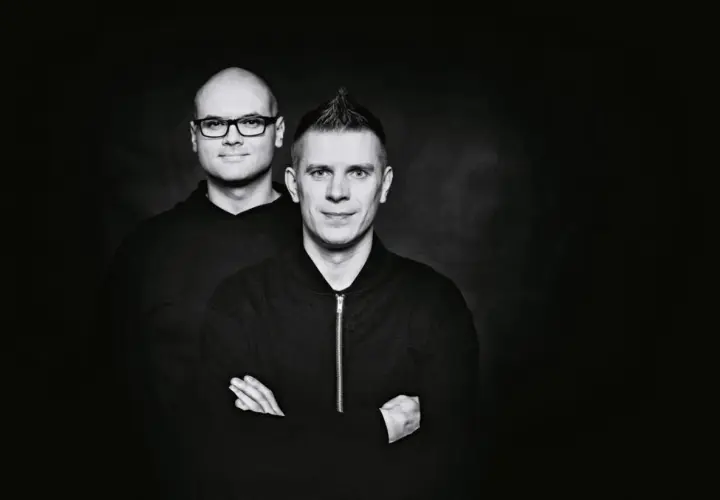 Dwóch mężczyzn w czarnych bluzach na ciemnym tle. Jeden z nich ma okulary, drugi irokeza. Wyglądają jakby właśnie wylądowali po locie dzika.