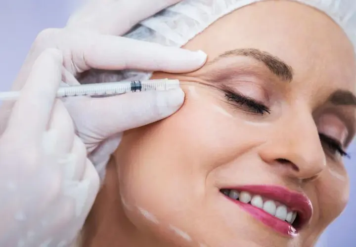 Kobieta uśmiecha się podczas zabiegu baby botox. Dłoń w rękawiczce trzyma strzykawkę z igłą, aplikując preparat w okolicę oka.