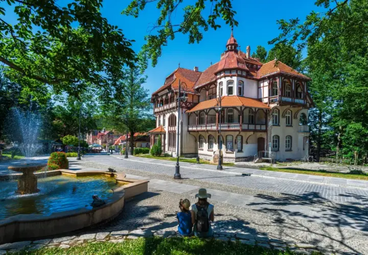 Dwie osoby siedzą przy fontannie, podziwiając architekturę luksusowego sanatorium w otoczeniu zieleni.