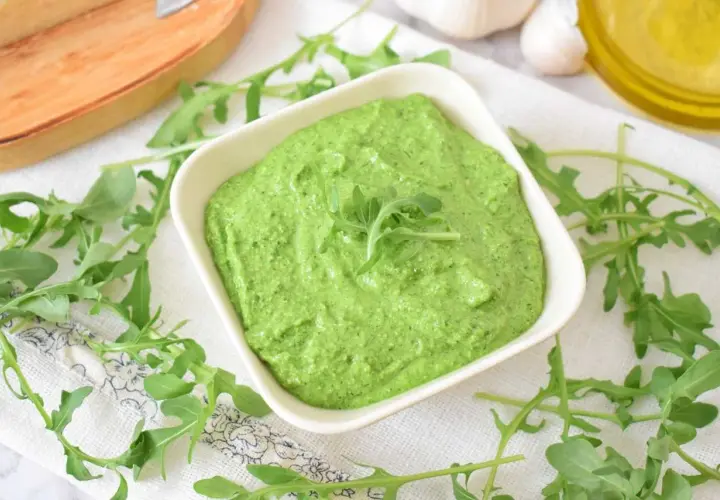 Aksamitne, zielone pesto z rukoli w białej miseczce, otoczone świeżymi listkami rukoli, czosnkiem i oliwą.