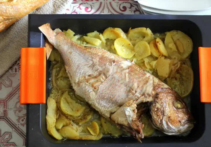 Pargo entero al horno con patatas y cebolla en bandeja negra con asas naranjas.