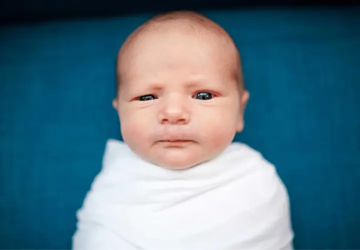 Un neonato senza capelli davanti, avvolto in una coperta bianca, guarda con occhi curiosi.