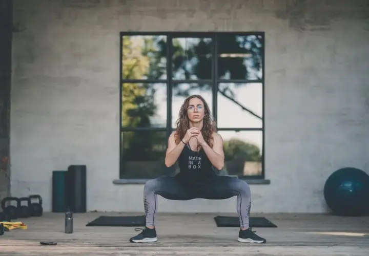 Kobieta wykonuje przysiad sumo. Widoczne hantle, piłka gimnastyczna i maty. Trening HIIT efekty widać po jej sylwetce.