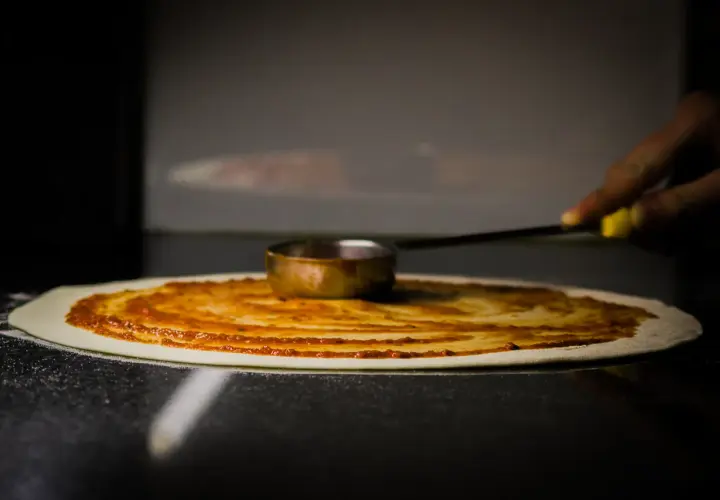Rozsmarowywanie sosu pomidorowego na cieście pizzy. To kluczowy etap leżakowania ciasta na pizzę, przygotowujący je do dalszych etapów.