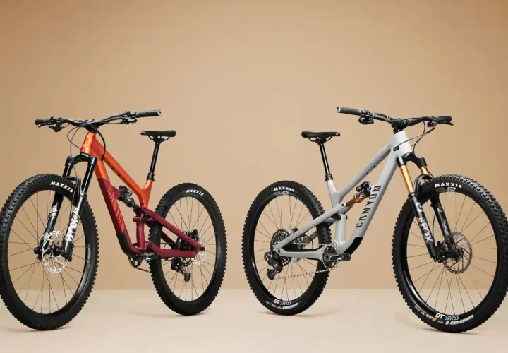 Two full-suspension mountain bikes, one orange and burgundy, the other grey, are shown side-by-side, highlighting the 27.5 vs 29 wheel size debate.