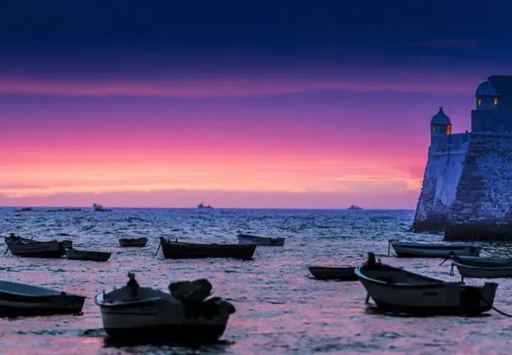 Barcas flotando en el mar al atardecer, con un castillo histórico y un cielo púrpura y rosa. Una hermosa puesta de sol en Cádiz.