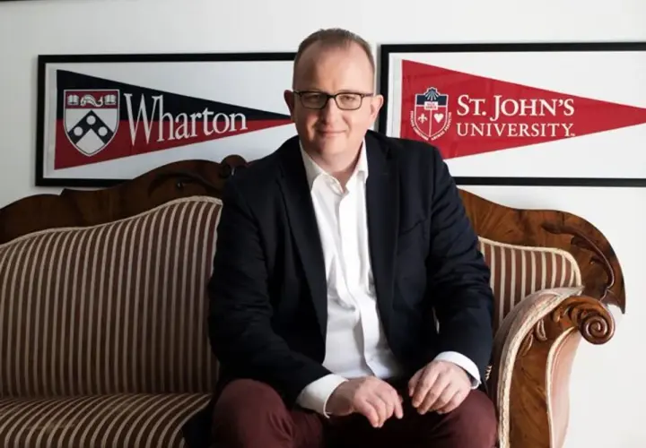Mężczyzna w garniturze siedzi na kanapie. W tle banery Wharton i St. John's University, prestiżowa uczelnia w USA.