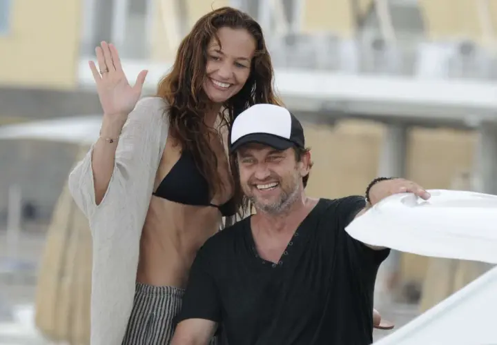 Gerard Butler z uśmiechniętą kobietą, która może być jego żoną, na plaży. Ona macha ręką, on ma na głowie czapkę.