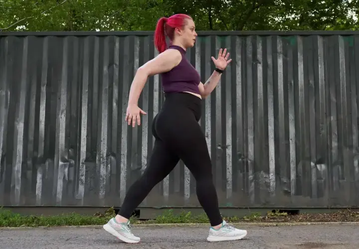 Kobieta z różowymi włosami w kucyku uprawia power walking przed szarym, pofałdowanym kontenerem.
