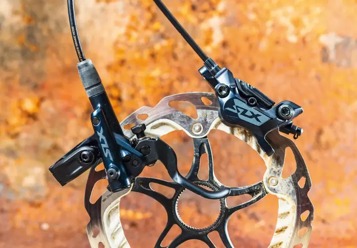 Shimano SLX M7120 hydraulic disc brakes, ready for any trail. The dark blue calipers and rotors are mounted on a rusty, textured background.