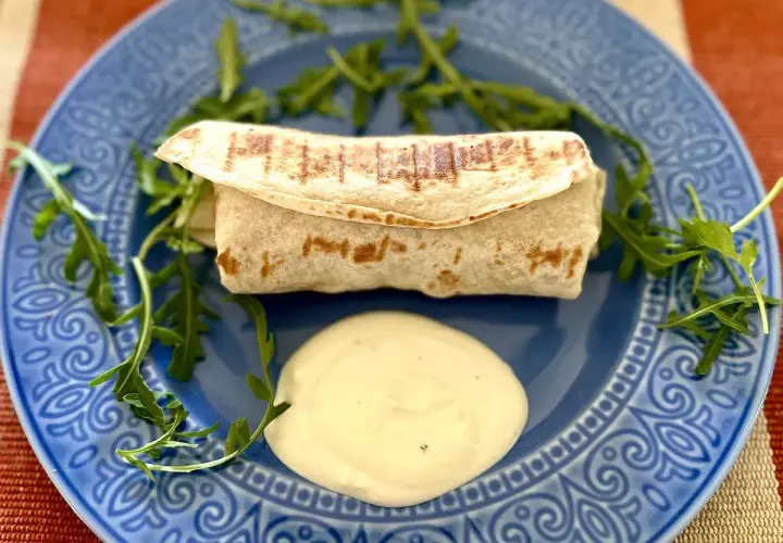 Ciepła tortilla z kurczakiem, podana z sosem i rukolą na niebieskim talerzu. Pyszne danie na szybki obiad.
